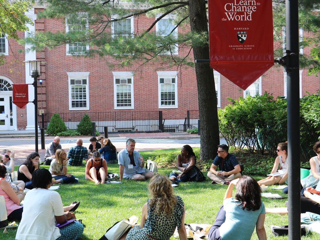 Participants sit in a circle on the lawn of Gutman Library in the summer, Longfellow Hall is in the foreground, a flag reads "Learn to Change the World"