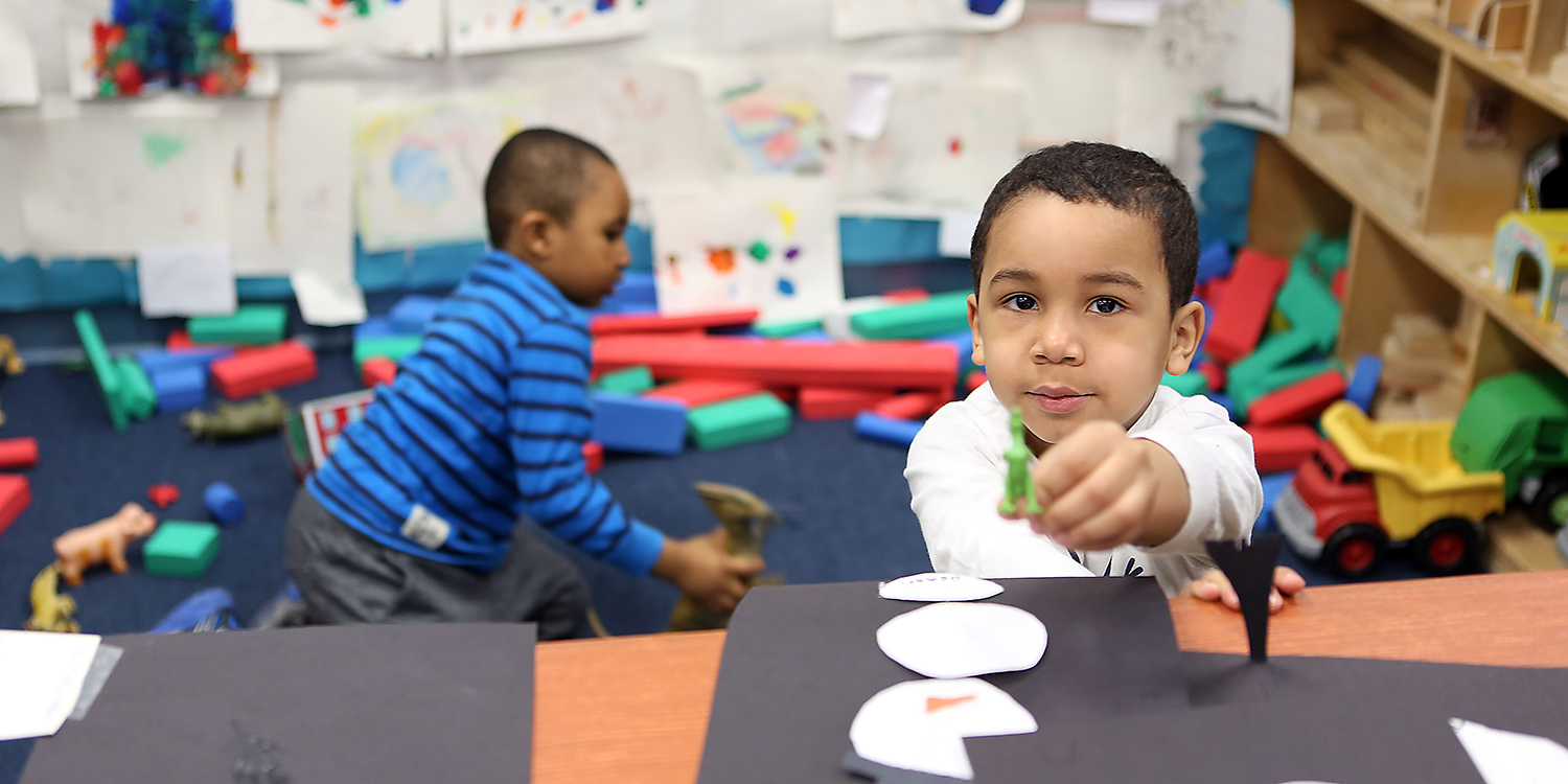 Preschool boy with art project, with another boy playing in the background