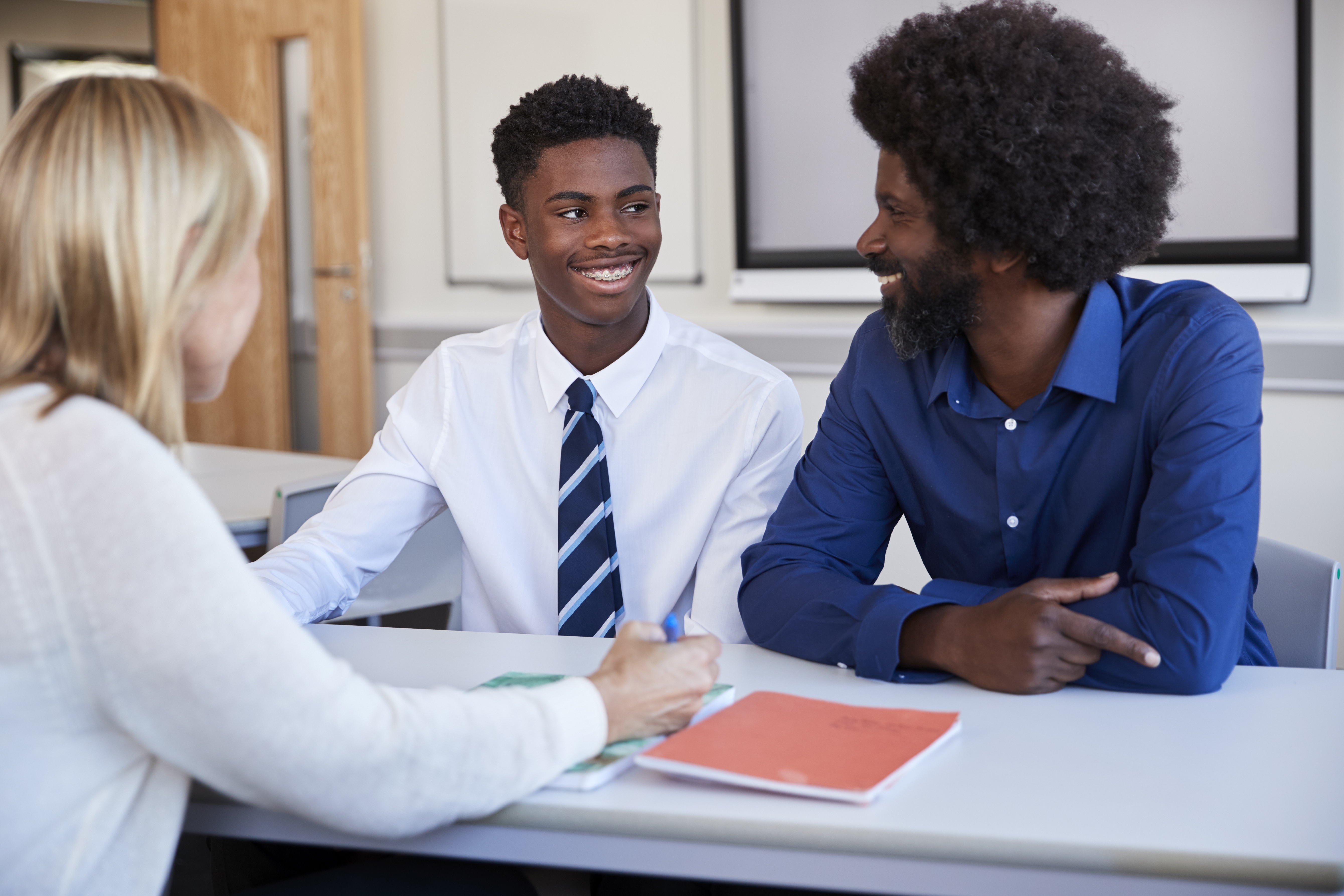 A black father and teenage son discuss with a female teacher at a high school parents' evening.