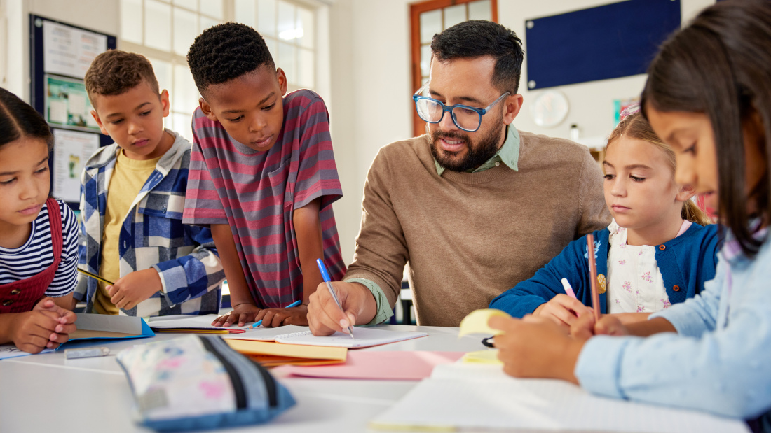 Five elementary school students, a mix of genders, gather around their male teacher, who is helping them with their work.