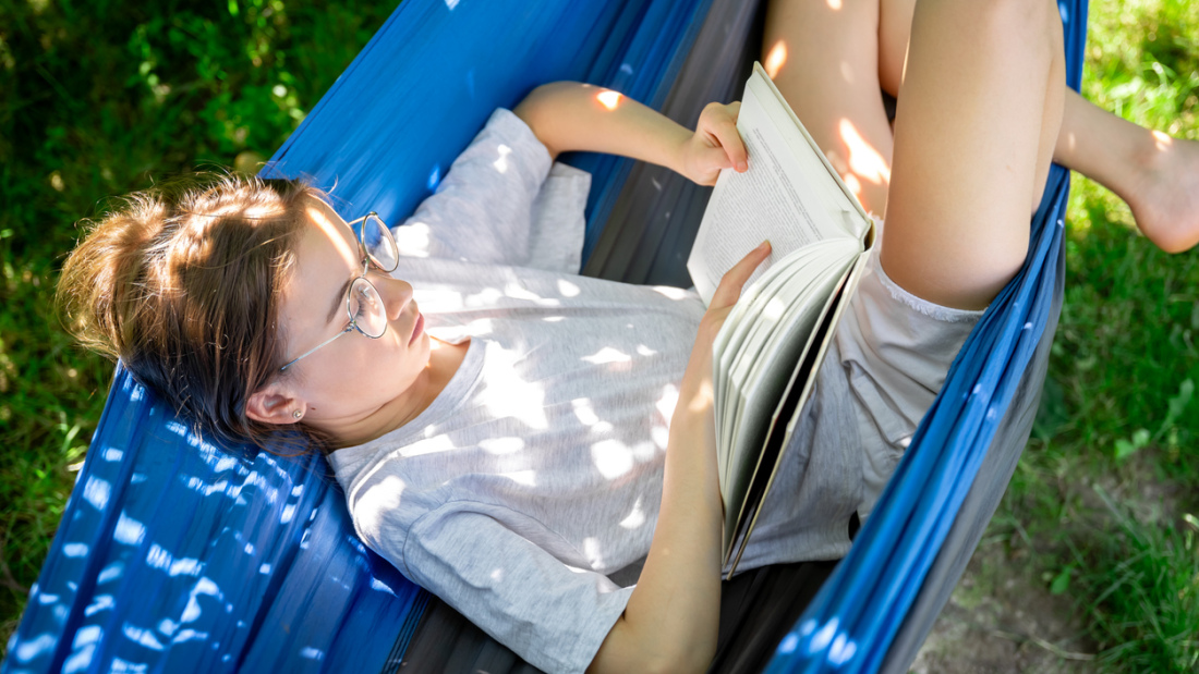 Photo of a preteen girl reading a book on a hammock during the summer