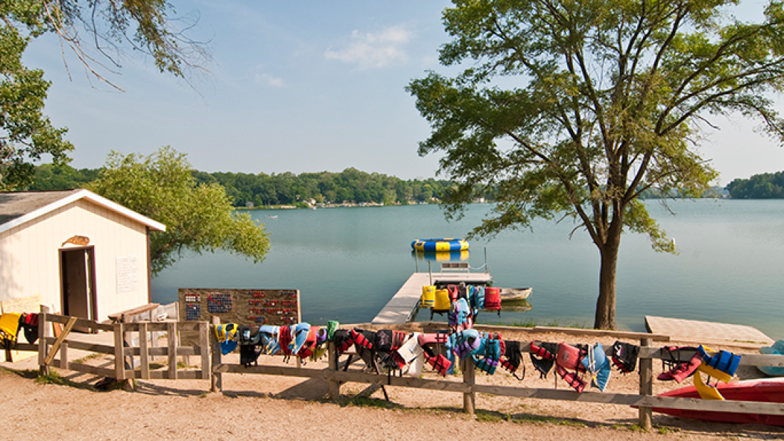 Picture of a lake in summertime, with a camp house and a wooden fence hung with colorful lifejackets 
