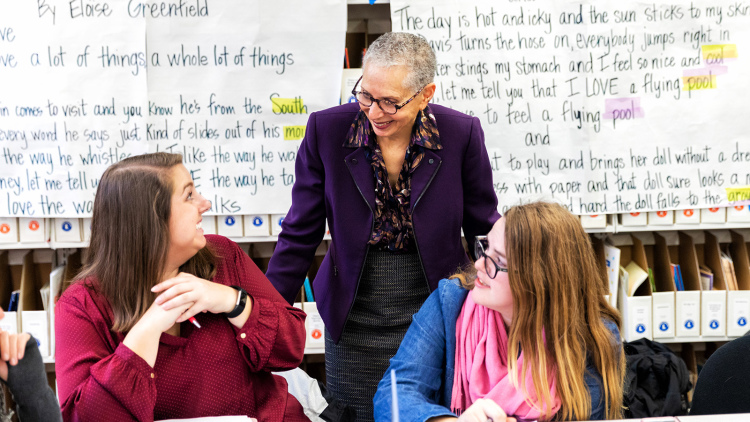 A faculty member interacts with two students in a classroom. In the background there are text-heavy sheets hanging on the walls.