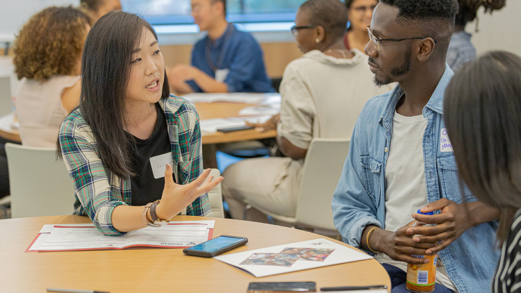 Students sit at a table with information packets. A young woman explains material