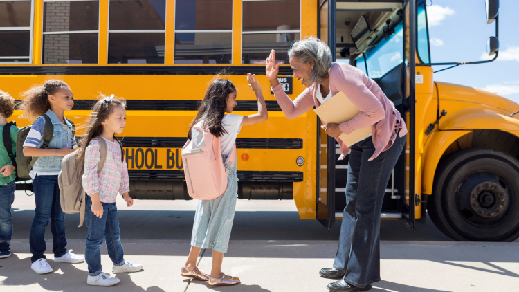 Adult woman educator high-fives elementary students as they get on the school bus.