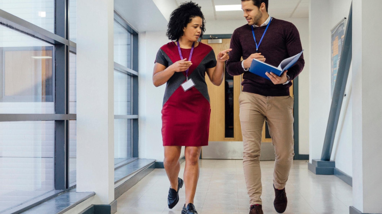 Two educators, a man and a woman, have a conversation walk down the hallway. 
