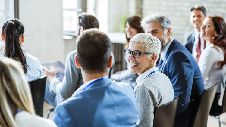 A large group of people on a seminar in a board room. The focus is on a smiling woman talking to her colleagues at a seminar.