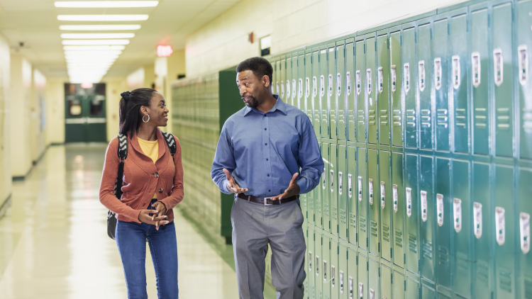 An African-American high school student walks with the principal in the hallway by a row of lockers. 