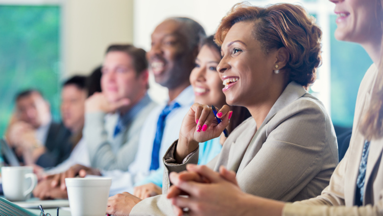 A black woman is smiling while attending professional development. She is dressed in smart business casual clothing and is sitting next to a diverse group of professional coworkers.