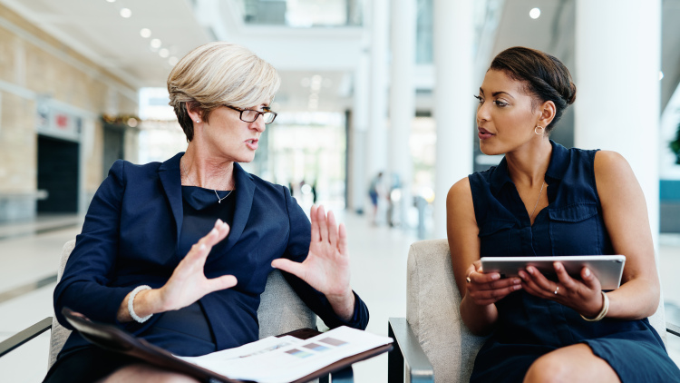 Two women having a discussion together while being seated inside an office building during the day.