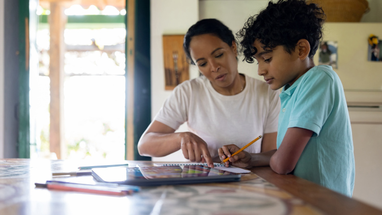A mother aiding her young son with homework at home. The boy focuses on writing while the mother prepares study materials.
