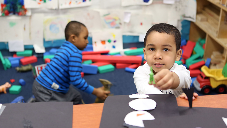 Preschool boy with art project, with another boy playing in the background