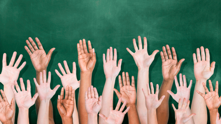 children's hands raised in front of a classroom chaulkboard