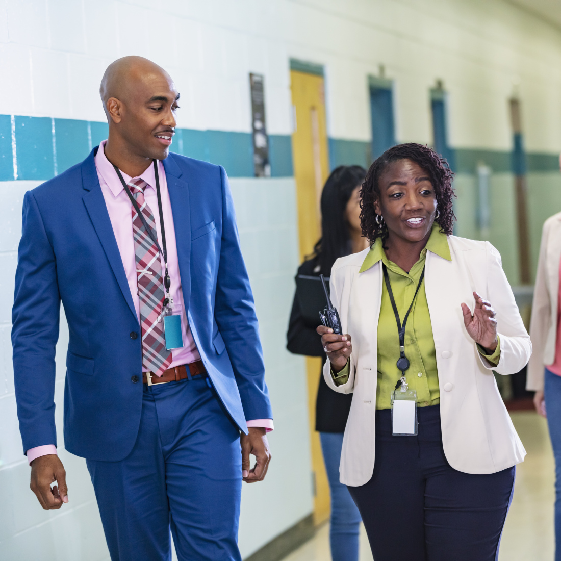 A multiracial group of five teachers or school administrators conversing as they walk through a school corridor. The focus is on the three in the foreground. The African-American woman in the middle, in her 50s, is talking while her coworkers look at her and listen.