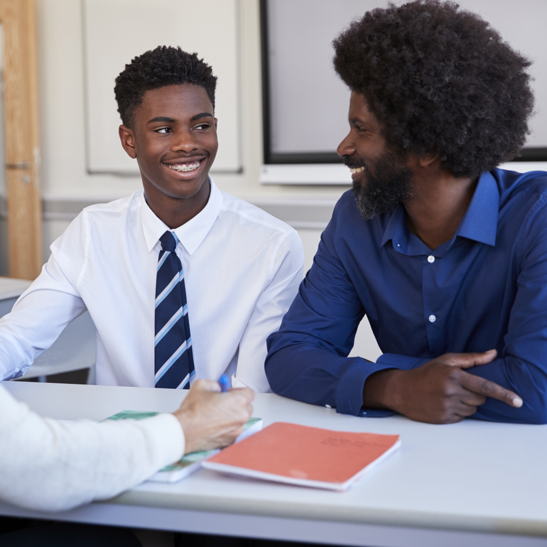 A black father and teenage son discuss with a female teacher at a high school parents' evening.