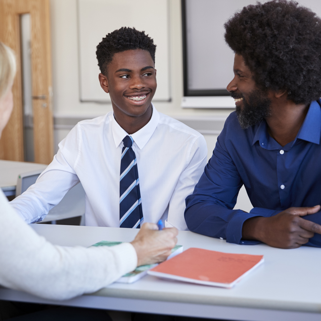 A black father and teenage son discuss with a female teacher at a high school parents' evening.