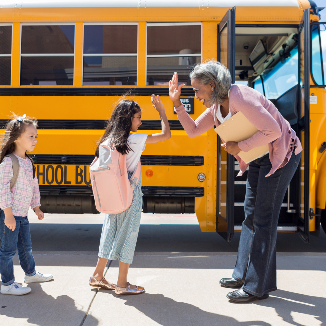 Adult woman educator high-fives elementary students as they get on the school bus.