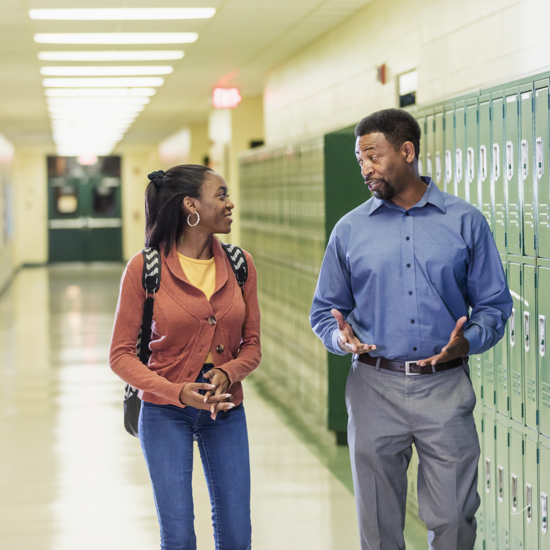 An African-American high school student walks with the principal in the hallway by a row of lockers. 