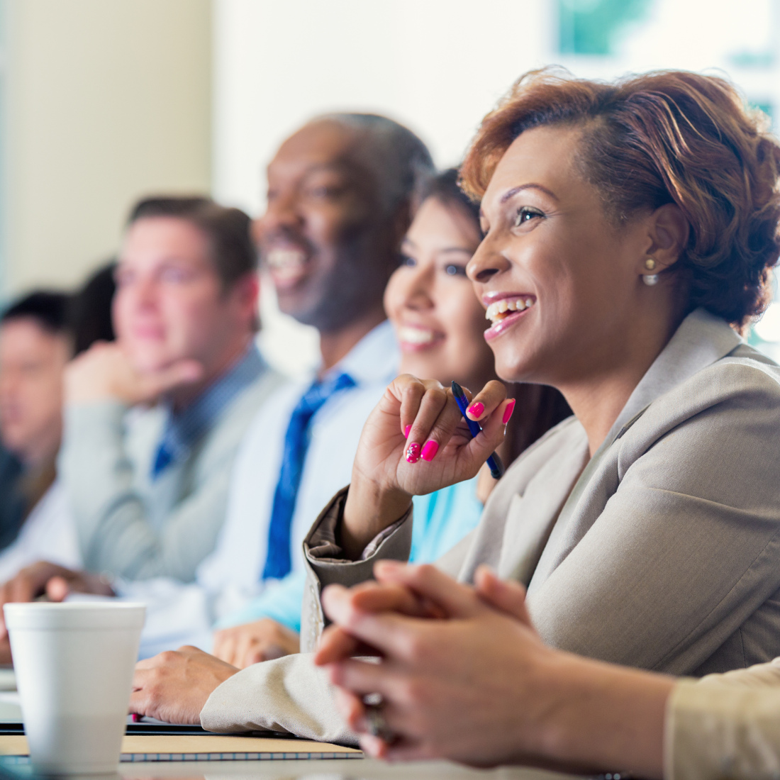 A black woman is smiling while attending professional development. She is dressed in smart business casual clothing and is sitting next to a diverse group of professional coworkers.