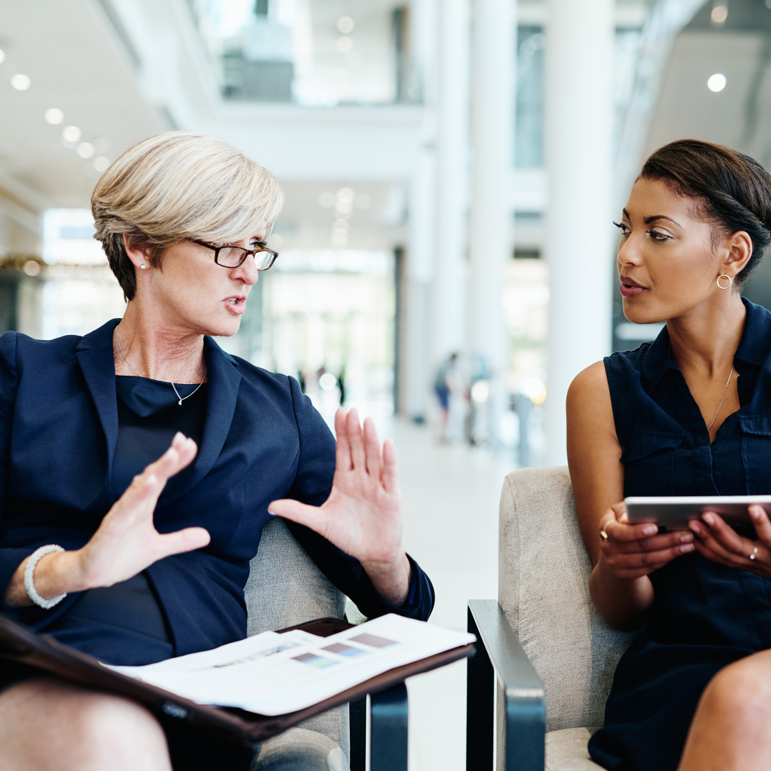 Two women having a discussion together while being seated inside an office building during the day.
