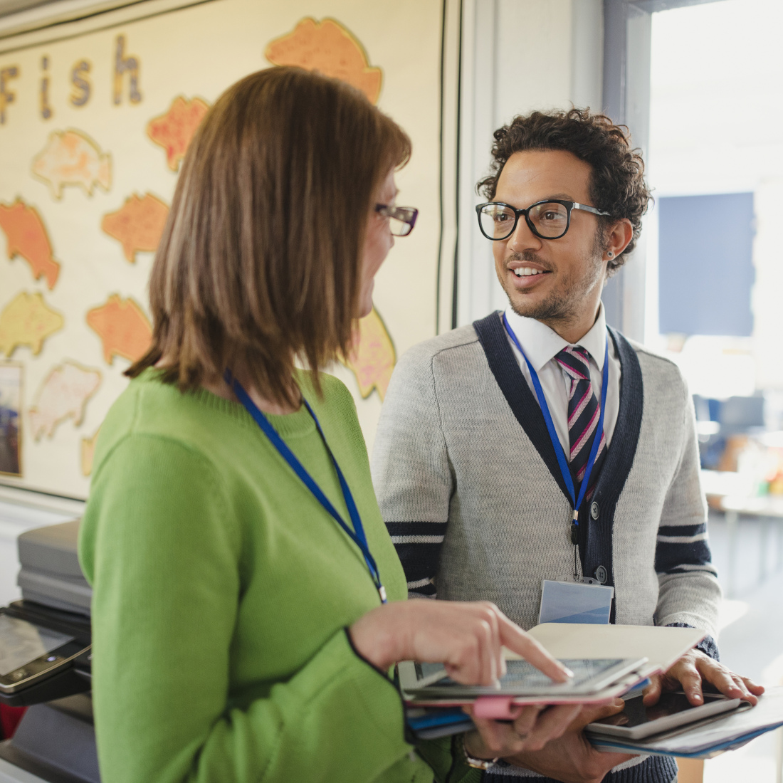 A principal discusses her work with her colleague; they are both carrying digital tablets and paper work.