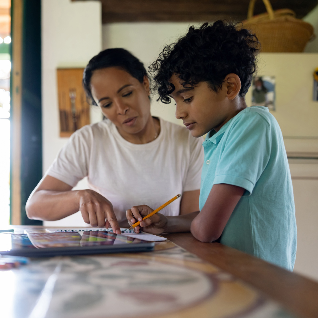 A mother aiding her young son with homework at home. The boy focuses on writing while the mother prepares study materials.