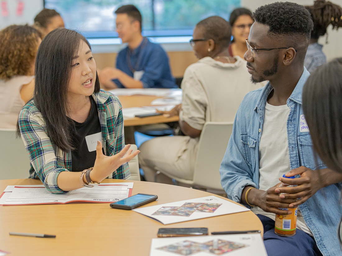 Students sit at a table with information packets. A young woman explains material