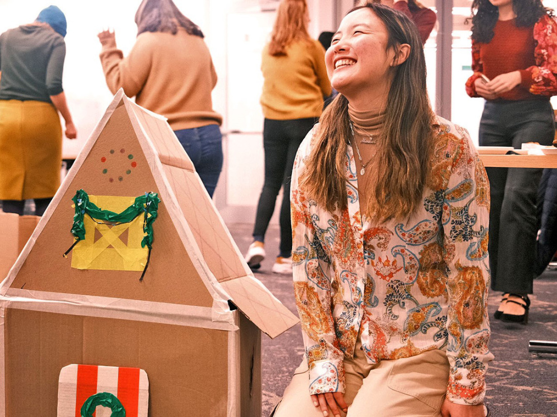 A student smiles while kneeling next to a cardboard structure art project.